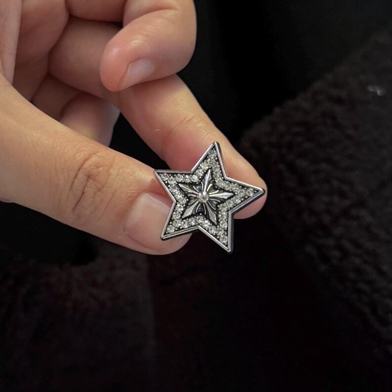 Op de afbeelding: Een zilveren stervormig accessoire met een centraal ontwerp en bezet met kleine, heldere edelstenen. De ster heeft een vijfhoekige vorm en wordt tegen een donkere achtergrond gehouden.