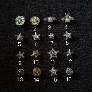Op de afbeelding: Een verzameling van zestien decoratieve knopen en spelden in verschillende vormen en ontwerpen. De collectie bevat ster-, bijen- en bloemmotieven, met versieringen zoals parels en strass. De knopen zijn in goud, zilver en parelkleuren.