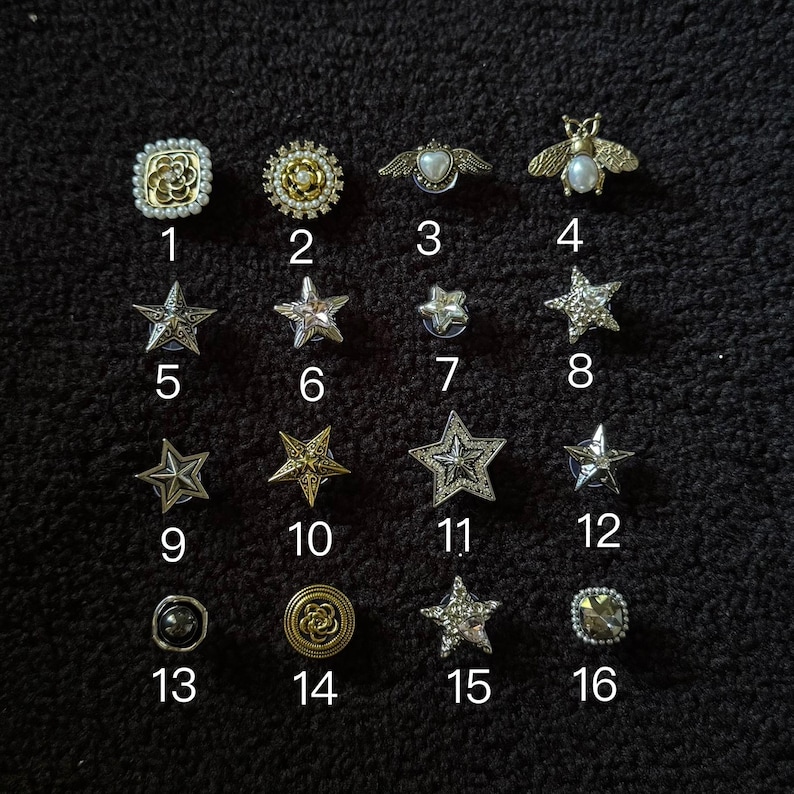 Op de afbeelding: Een verzameling van zestien decoratieve knopen en spelden in verschillende vormen en ontwerpen. De collectie bevat ster-, bijen- en bloemmotieven, met versieringen zoals parels en strass. De knopen zijn in goud, zilver en parelkleuren.
