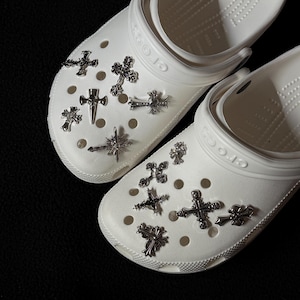 May include: A pair of white clogs adorned with various silver cross charms. The crosses are of different sizes and designs, adding a unique, personalized touch to the footwear. The clogs are made of a smooth, rubbery material.