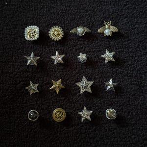 Op de afbeelding: Een verzameling decoratieve spelden en broches in verschillende vormen, waaronder sterren, bijen en bloemmotieven. De spelden hebben zilveren, gouden en parelaccenten. De spelden worden gepresenteerd op een donkere, getextureerde ondergrond.