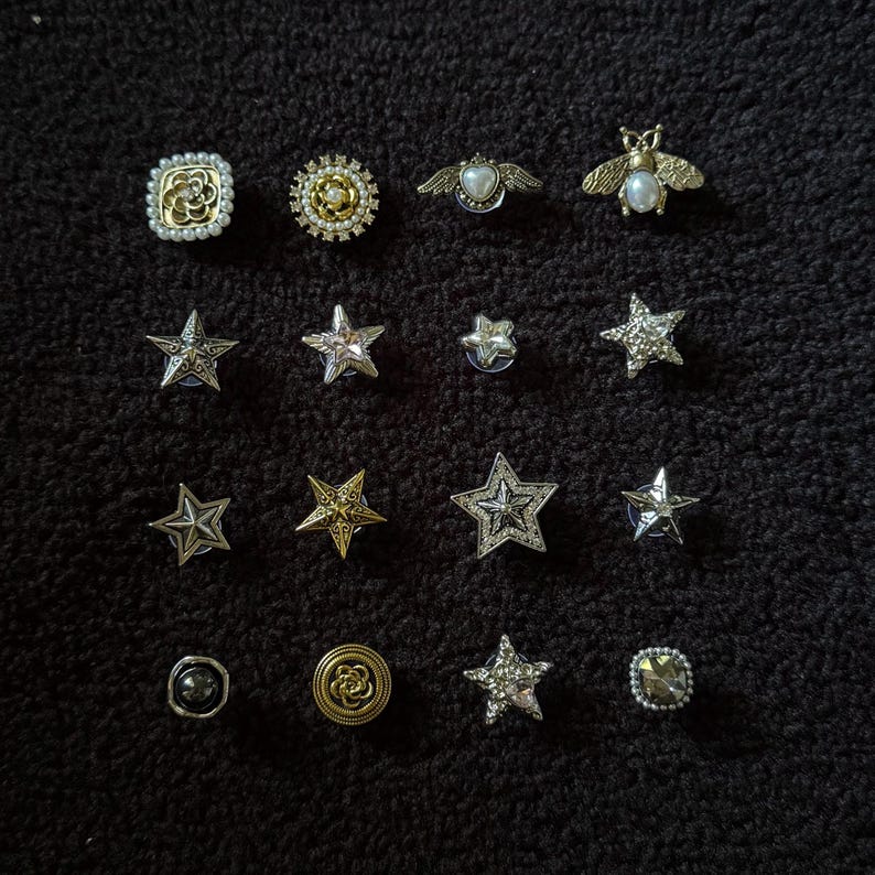 Op de afbeelding: Een verzameling decoratieve spelden en broches in verschillende vormen, waaronder sterren, bijen en bloemmotieven. De spelden hebben zilveren, gouden en parelaccenten. De spelden worden gepresenteerd op een donkere, getextureerde ondergrond.