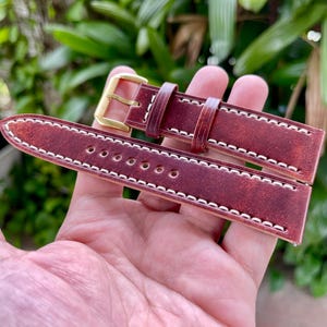 May include: A burgundy leather watch strap with white stitching and a gold-colored buckle. The strap is held in a hand, showcasing the detailed stitching and the rich color of the leather. The strap is designed for a watch.
