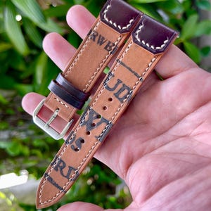 May include: Two-piece watch strap in brown leather with dark burgundy accents. The strap features white stitching and stamped text. A silver buckle is visible on the left side. The background is a blurred view of green foliage.