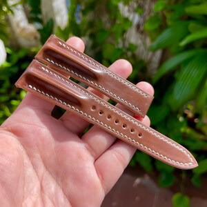 May include: A brown leather watch strap with white stitching. The strap has a gradient color effect, transitioning from a lighter shade to a darker brown. The strap is held in a hand against a blurred green background.