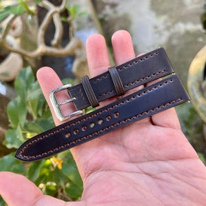 May include: A dark brown leather watch strap with contrasting stitching. The strap features a silver buckle and multiple adjustment holes. The leather has a smooth finish and is designed for a classic timepiece. The strap is shown against a blurred background.