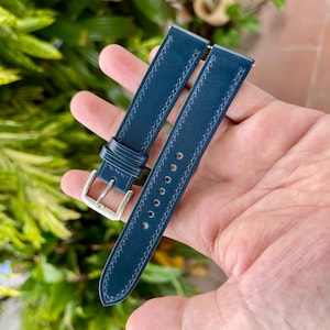 May include: A dark blue leather watch strap featuring tonal stitching along the edges, a silver-toned metal buckle, and a series of circular adjustment holes. The strap consists of two pieces, one with the buckle and one with the holes.