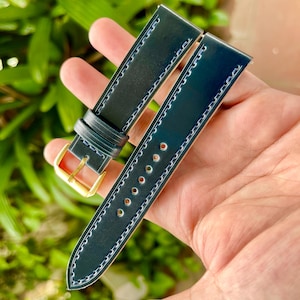 May include: Dark teal watch strap with white stitching and a gold buckle. The leather strap has a glossy finish and is held in a hand against a blurred green background. The strap has multiple holes for adjustment.