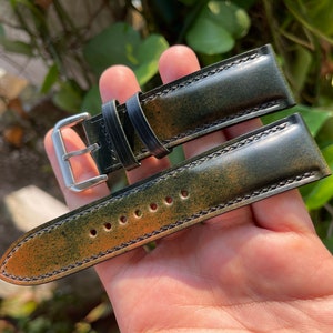 May include: A green leather watch strap with a brown patina and black stitching. The strap has a silver buckle.