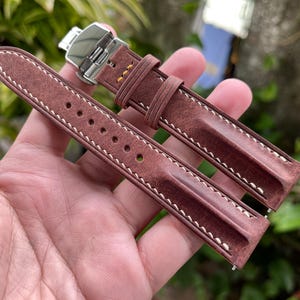 Puede incluir: Una correa de reloj de cuero marrón con costuras blancas y un cierre plateado. La correa tiene un aspecto texturizado y varios agujeros para el ajuste. La correa del reloj es de un color marrón rico y cálido.