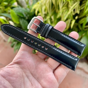May include: A black watch strap with a silver buckle. The strap is made of a glossy material and has a stitched edge. The strap is held in a person's hand against a background of green foliage.