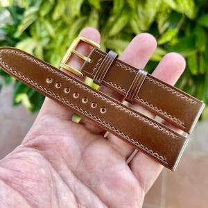 May include: A brown leather watch strap with a gold-colored buckle. The strap has white stitching along the edges and multiple holes for adjustment. The watch band is a rich, warm brown color.
