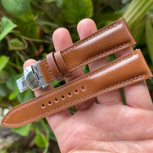 Pode incluir: Uma pulseira de relógio de couro marrom com uma fivela de prata. A pulseira tem um padrão costurado e é mostrada em uma mão.