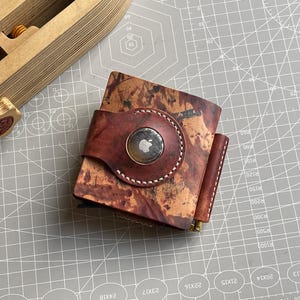 Op de afbeelding: Een bruine leren portemonnee met een uniek gemarmerd patroon. De portemonnee heeft een ronde houder voor een Apple AirTag en een contrasterende bruine leren riem. De stiksels zijn wit. De portemonnee ligt op een grijze ondergrond.