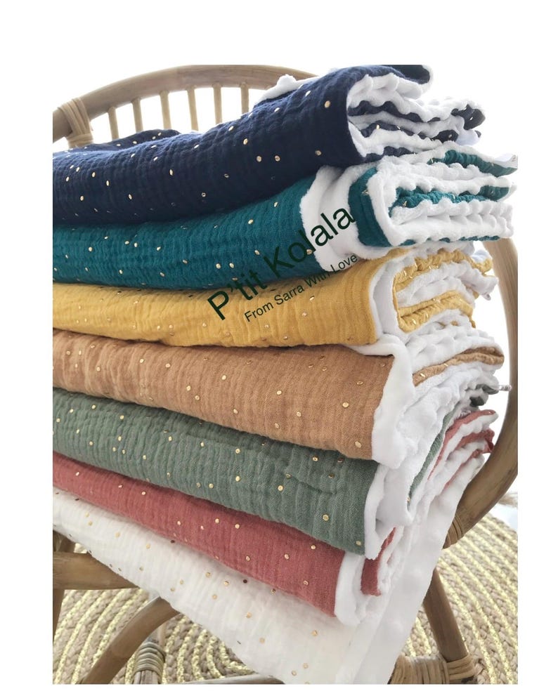 Op de afbeelding: Een stapel van zes babydekens in verschillende kleuren, waaronder wit, roze, groen, bruin, geel en blauw. Elke deken heeft een witte rand en gouden stippen. De dekens zijn gevouwen en gestapeld op een houten stoel. De tekst "Petit Koala From Sarra With Love" is zichtbaar op de gele deken.