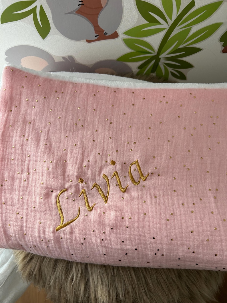 Op de afbeelding: Een roze babydeken met gouden glitter stippen en de naam "Livia" geborduurd in gouden draad.