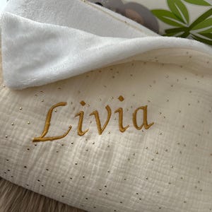 Op de afbeelding: Een wit en gouden babydeken met een gouden geborduurde naam "Livia" erop.