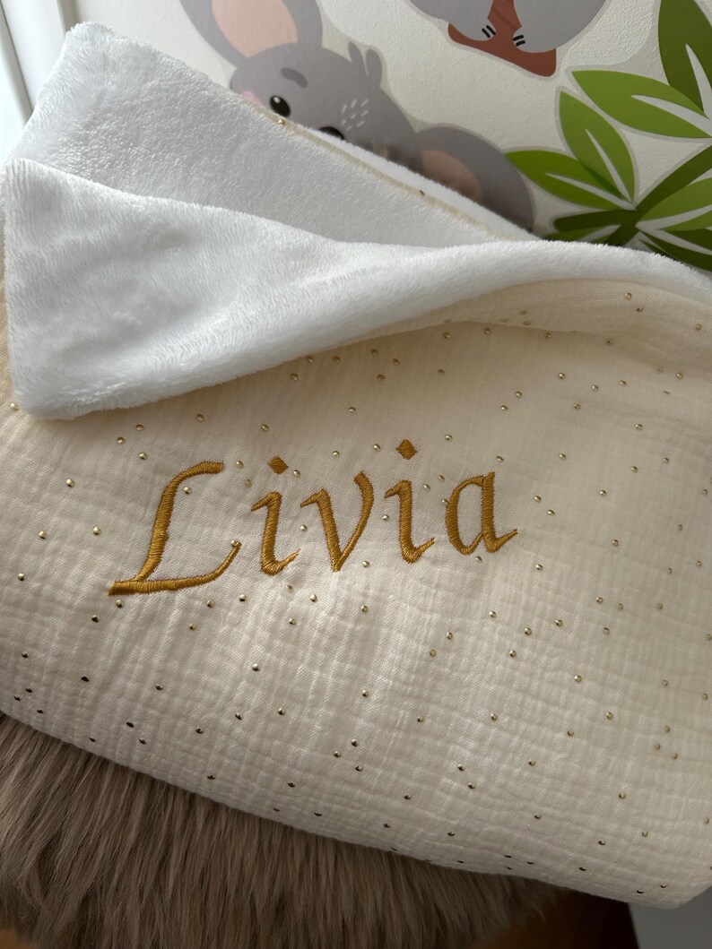 Op de afbeelding: Een wit en gouden babydeken met een gouden geborduurde naam "Livia" erop.