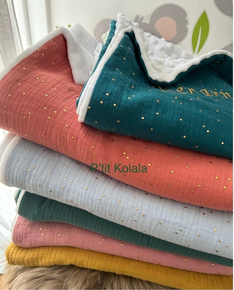 Op de afbeelding: Een stapel van zes zachte, katoenen babydekens in verschillende kleuren, waaronder koraal, teal, lichtblauw, roze en geel. Elke deken heeft een subtiel patroon van gouden stippen en een witte rand. De bovenste deken heeft de tekst "P'tit Kolala" geborduurd in goud.