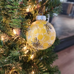 May include: A yellow Christmas ornament with a honeycomb pattern and black bee illustrations. The ornament is hanging on a green Christmas tree with small white lights. The ornament is round and has a silver cap.