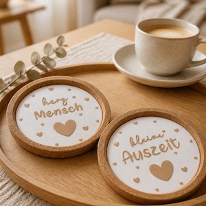 May include: Two round coasters with a cork border and white center. One coaster has the text "herz Mensch" and a heart, the other says "kleine Auszeit" with a heart. A cup of coffee sits on a saucer on a wooden tray.