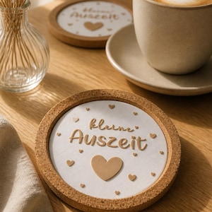 May include: Cork coasters with a white center and brown border. The coasters have the words "kleine Auszeit" and heart shapes. A cup of coffee sits on a saucer next to a coaster.