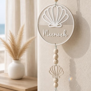 May include: A circular, beige wall decoration featuring a seashell design and the word "Meerweh". It's suspended from a wooden peg with a string and wooden bead. Below, a seashell shape and wooden beads lead to a clear crystal pendant.