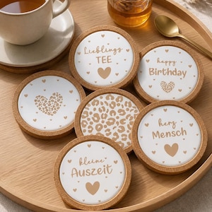 May include: A set of six round coasters with cork rims, featuring white centers with various designs and text in a light brown color. Designs include hearts, leopard print, and phrases like "happy Birthday". A cup of tea and honey are also present.