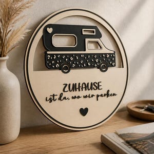 Könnte beinhalten: Runder Holzschild mit einem schwarz-weißen Wohnmobil-Design mit Leopardenmuster. Der deutsche Text lautet "ZUHAUSE ist da, wo wir parken". Enthält ein Herzsymbol. Wohnaccessoire.