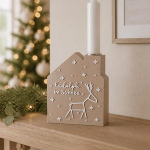 May include: A light brown house-shaped candle holder with a white taper candle inserted in the top. The front features a white line drawing of a reindeer, the text "Rudolph im Schnee," and several small white star-shaped accents scattered across.