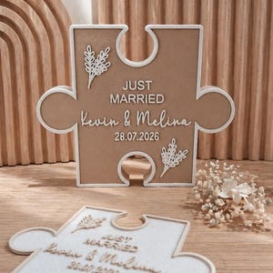 Op de afbeelding: Een beige en wit puzzelstuk met de woorden "JUST MARRIED" en de namen "Kevin & Melina" samen met de datum "28.07.2026". Het puzzelstuk heeft een witte rand en decoratieve bloemaccenten. Bruiloft decoratie.