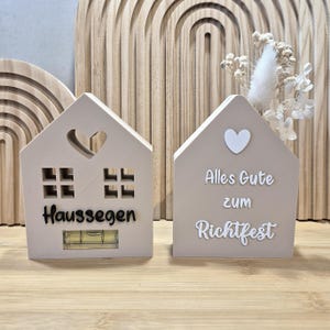 Op de afbeelding: Twee beige houten huisvormige decoraties. Eén heeft een hartuitsparing en het woord "Haussegen" met een waterpas. De andere heeft een wit hart en de woorden "Alles Gute zum Richtfest". Gedroogde bloemen op de achtergrond.