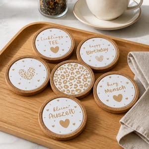 May include: A collection of six round coasters with cork borders and white centres. Each coaster displays a unique design, including text such as "Happy Birthday" and "Lieblings TEE", hearts, and a leopard print. A cup of tea and a jar of tea leaves are also present.