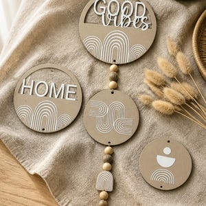 Puede incluir: Un adorno de pared decorativo con cuatro placas de madera circulares. La placa superior dice "GOOD VIBES" en escritura blanca. Otras placas presentan diseños geométricos blancos. El colgante está adornado con cuentas de madera y un cristal.