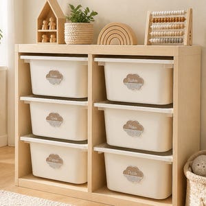 May include: A light wood toy storage unit with six white plastic bins, each featuring a cloud-shaped label with the text Bouen, Basteln, Spiele, Malen, Puppen, and Puzzle. The top surface holds a wooden house, plant, rainbow, and abacus.