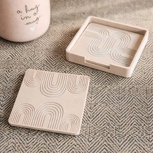 May include: Beige square coasters with a carved rainbow design. One coaster is separate, and the other is in a matching tray. A pink mug with the words "a hug in a mug" is in the background.