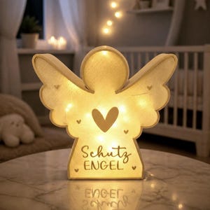 May include: A light-up angel-shaped wooden ornament with a warm glow, featuring a central heart cutout, smaller heart accents, and the text "Schutz ENGEL" printed on the front. The piece emits a soft light from within its hollow structure.