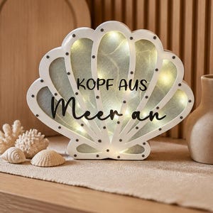 Peut inclure: Une lumière en forme de coquillage blanc avec le texte allemand "Kopf aus Meer an" en écriture noire. La lumière contient de petites ampoules de couleur chaude. Des coquillages et du corail décoratifs sont disposés devant la lumière.