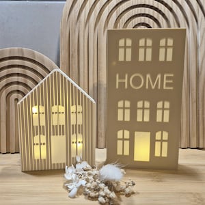Puede incluir: Dos lámparas decorativas con forma de casa. Una es una casa con rayas verticales, y la otra es un edificio alto con la palabra "HOME" recortada. Ambas tienen ventanas iluminadas. Flores secas están frente a las casas.