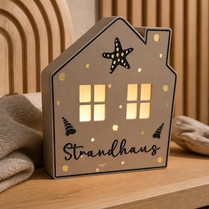 Peut inclure: Lampe décorative en forme de maison avec une étoile de mer et le mot "Strandhaus". La lampe a deux fenêtres éclairées et est décorée de points dorés et de motifs de coquillages. Elle est posée sur une surface en bois.