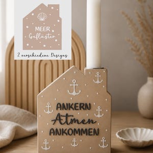 May include: A beige house-shaped candle holder with white anchor designs and the words "ANKERN, Atmen, ANKOMMEN" in black. A white candle sits in the holder. A small card with the text "MEER, Geflüster" is in the background.