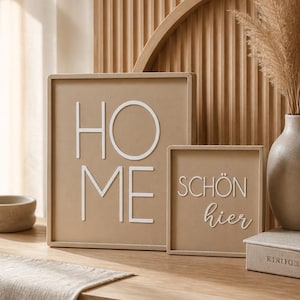 Può includere: Due quadri da parete incorniciati con le parole "HOME" e "SCHÖN hier" in bianco, su sfondo beige. L'arredamento è posto su una superficie di legno, con un vaso ed elementi decorativi.
