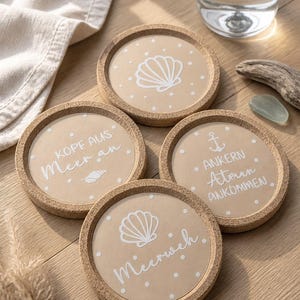 May include: Set of four round cork coasters with a beach theme. Each coaster features a beige background with a white seashell design or text. The text includes phrases like "Kopf aus Meer an" and "Meerweh."