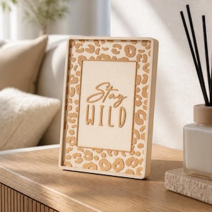 May include: A rectangular picture frame with a light beige border and a tan leopard print pattern. The centre features a cream-coloured insert with the text Stay WILD in raised, tan-coloured lettering, presented in a clean, upright orientation.