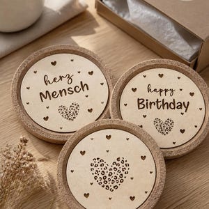 Könnte beinhalten: Drei runde Untersetzer mit Korkrändern und hellen Holzmitteln. Einer der Untersetzer trägt die Aufschrift "herz Mensch", ein anderer "happy Birthday", und der dritte hat ein Leopardenherz. Kleine Herzdesigns umgeben den Text und das Leopardenherz.