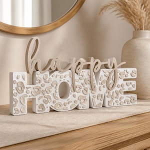 Könnte beinhalten: Dekoratives Schild mit der Aufschrift "happy HOME". Das Wort "HOME" ist in Blockbuchstaben mit weißem Grund und einem beigefarbenen Leopardenmuster gehalten. Das Wort "happy" ist kursiv und beige. Das Schild steht auf einer Holzoberfläche.