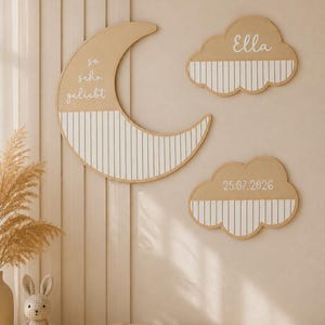 Könnte beinhalten: Ein Set aus drei Wanddekorationen aus Holz, bestehend aus einer Mondsichel und zwei Wolken. Der Mond ist mit dem Text "so sehr geliebt" beschriftet. Eine Wolke zeigt den Namen "Ella" und die andere das Datum "25.07.2026". Jedes Teil hat eine natürliche Holzoberfläche mit weißen vertikalen Streifen.