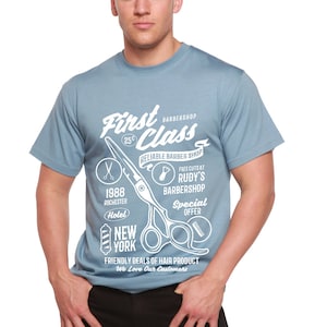 Puede incluir: Una camiseta azul claro con un diseño de barbería. El gráfico blanco incluye las palabras "First Class Barbershop", un par de tijeras y otro texto. La camiseta se usa con pantalones negros.
