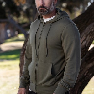 Puede incluir: Una sudadera con capucha verde con cremallera, capucha y cordón. La sudadera tiene un bolsillo canguro y puños y cintura de canalé.