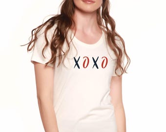 May include: White t-shirt with a scoop neck and short sleeves. The shirt features the letters "XOXO" in navy blue and red. The model is wearing black pants.
